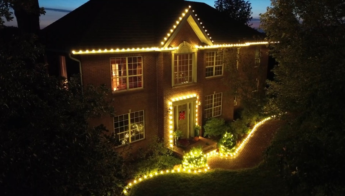 Two-story brick home in Lincoln, Nebraska lit with warm white Christmas lights along the roofline, around the front door and down the curved walkway at dusk